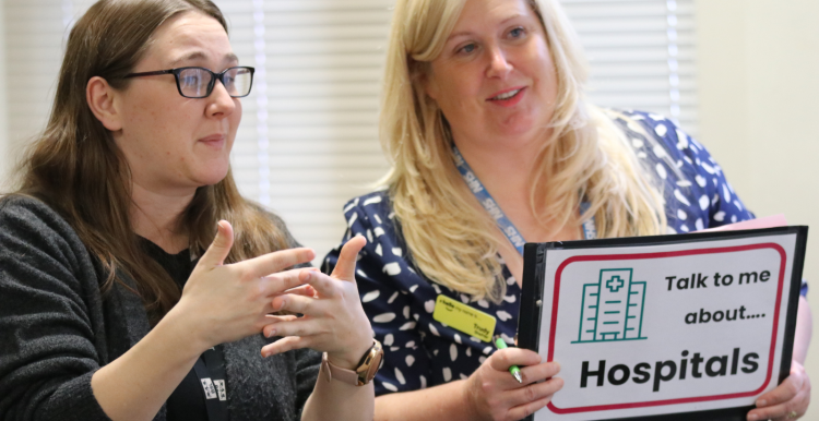 A woman holding up a sign saying 'Talk to me about Hospitals' and a woman next to her signing.