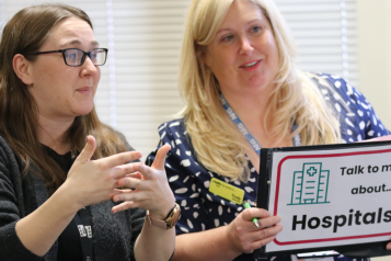 A woman holding up a sign saying 'Talk to me about Hospitals' and a woman next to her signing.